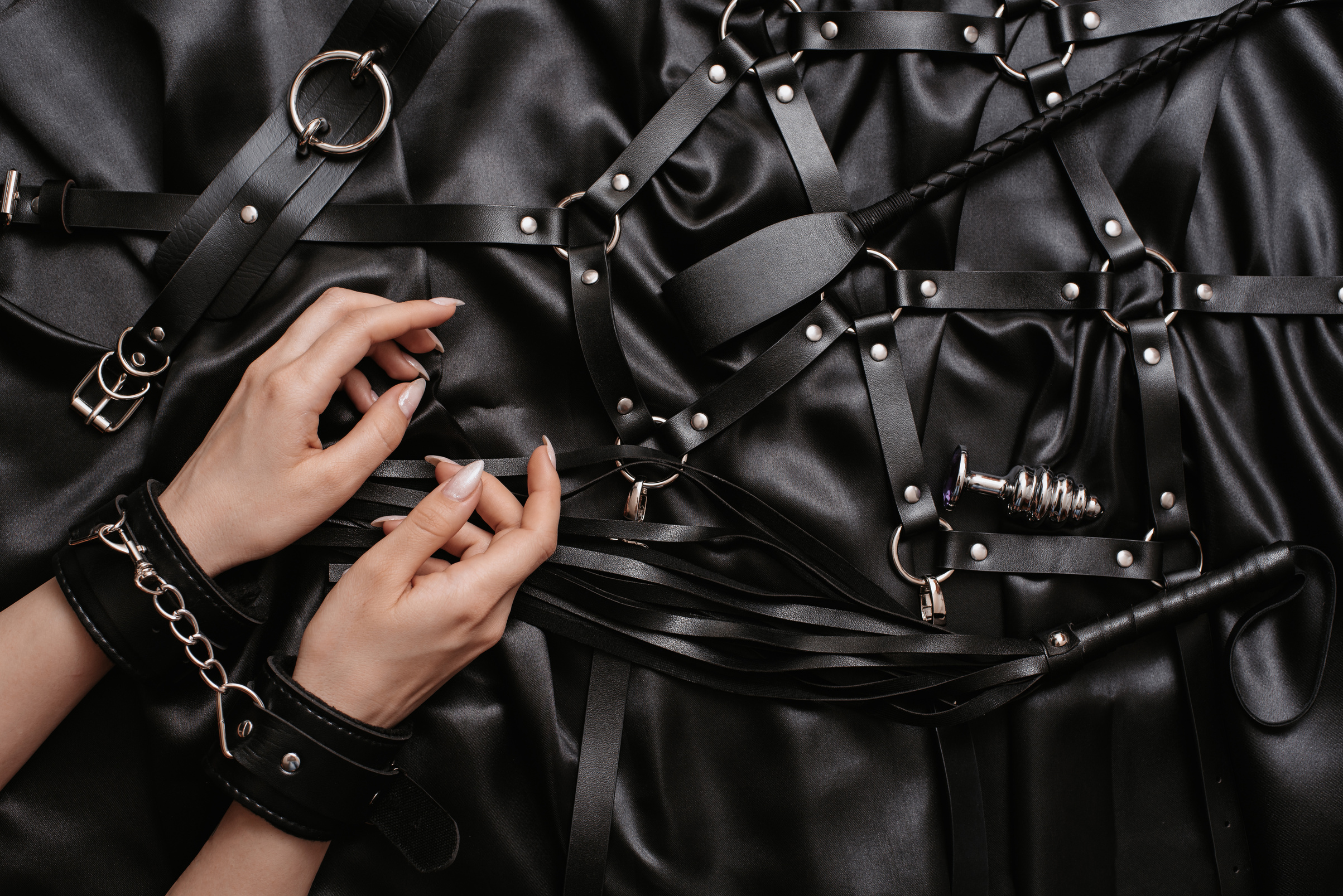 Women's Hands in Handcuffs on a Dark Silk Sheet Next to Adult Toys